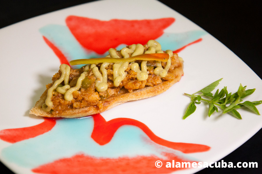 Bruschetta con enchilado de pescado cubierto con un espejo de salsa guacamole decorado con un guinda de pimiento encurtido.